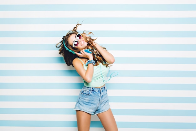 Free Photo expressing brightful positive emotions of young joyful woman in moving listening to music throgh headphones on striped wall. summer look, long curly brunette hair, sunglasses, true emotions.