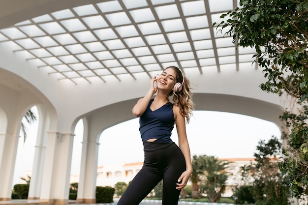 Free photo expressing positivity of joyful pretty woman in sportswear by training smiling on east architecture. listening to music through headphones, happiness, sportive lifestyle