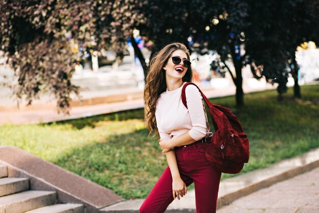 Expressive girl with long curly hair is posing with vinous bag in park in the city. She wears marsala color, sunglasses and has good mood.