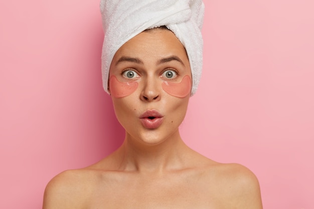 Free Photo under eye treatment and body care concept. shocked young caucasian woman applies beauty patches, removes dark circles and puffiness, looks surprisingly at herself in mirror, stands topless indoor