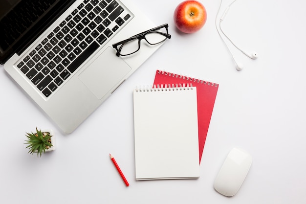 Eyeglasses on laptop,apple,earphones,colored pencil,spiral notepad and mouse on white desk