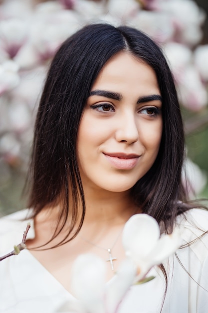 Face portrait of beautiful caucasian brunette girl near blossom magnolia tree outdoors in spring park.