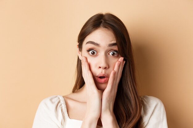 Face of surprised girl saying wow touching cheeks and staring excited at camera standing on beige ba...