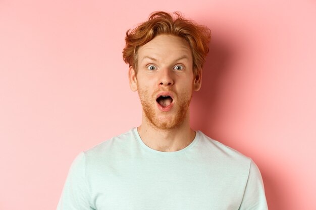 Face of surprised redhead man reacting to cool promo offer, raising eyebrows and gasping, staring in awe at camera, standing over pink background.