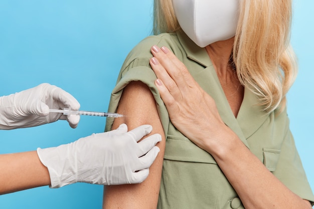 Faceless blonde woman wears protective face mask dress gets inoculaton in shoulder for disease prevention poses against blue wall