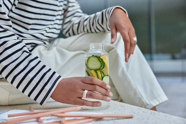 Free Photo faceless woman in casual clothes holds bottle of detox drink water with sliced lemon and cucumber rests aftr drawing picture surrounded by colored pencils