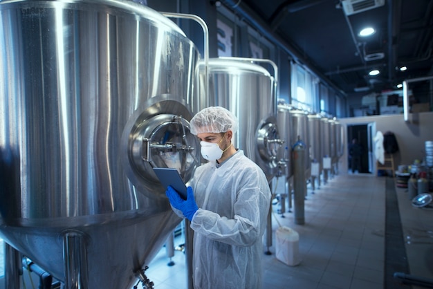 Factory worker technologist in protective uniform with hairnet and mask controlling food production on tablet computer