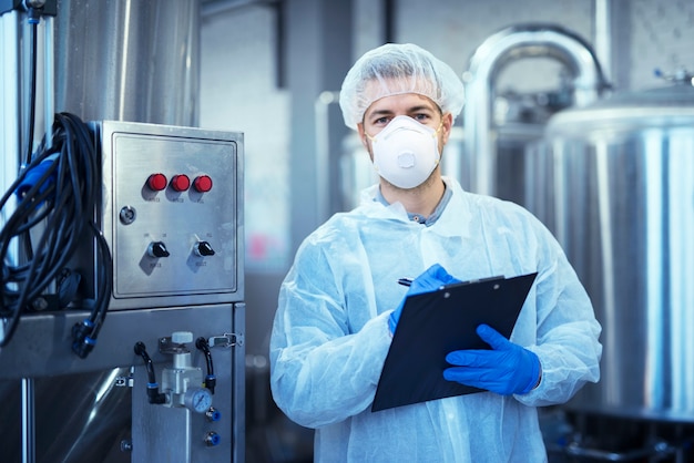 Factory worker in white protective uniform with hairnet and mask standing by industrial machine