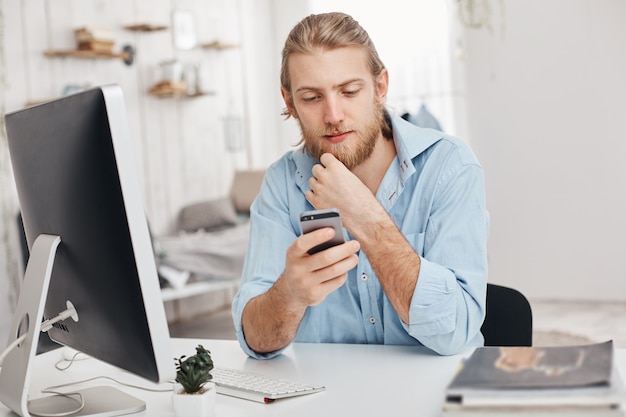 Free Photo fair-haired bearded male freelancer installs new app on smart phone, downloads program on computer, uses wi-fi, recieves message from partner. business, modern technologies, communication