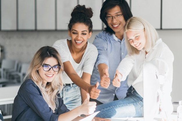 Fair-haired female it-specialist fooling around with friends, sitting at workplace and laughing. Excited japanese manager posing with smile, standing beside blonde colleague in office.