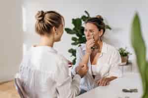 Free photo fair-skinned adult woman uses roller massager while sitting next to her beautician in light studio. home spa facial concept