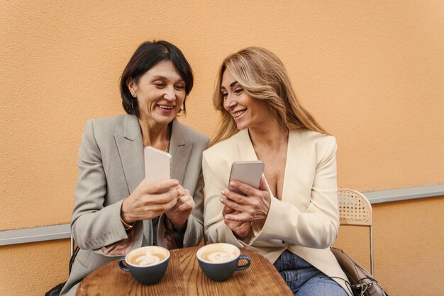 Fairskinned adult sisters in casual clothes use smartphones and drink coffee in street cafe Family and technology concept