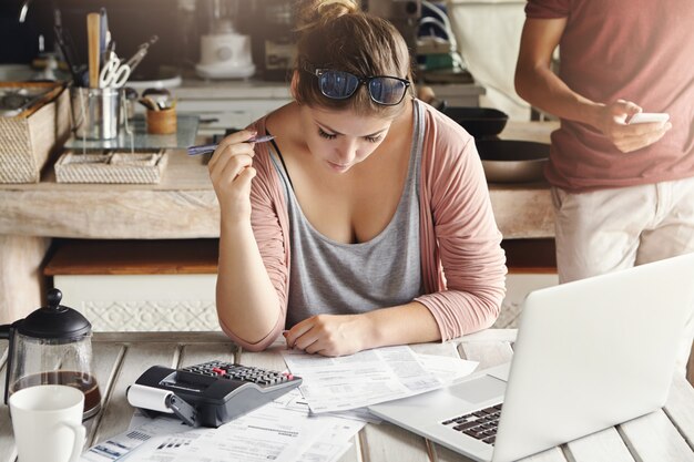 Family budget and financial problems. Concentrated worried woman doing paperwork at home, calculating domestic expenses and paying gas and electricity bills, using laptop computer and calculator