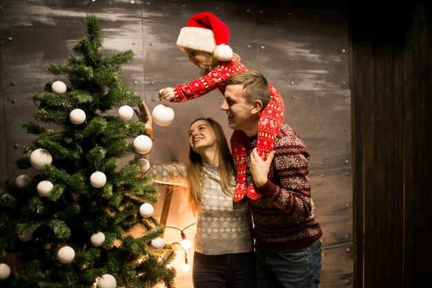 Family by the Christmas tree with little daughter in a red hat