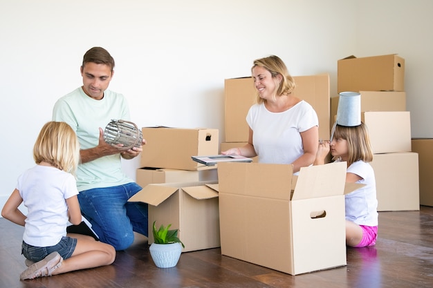 Free photo family couple and little girls moving into new flat, having fun while unpacking things in new apartment, sitting on floor and taking objects from open boxes