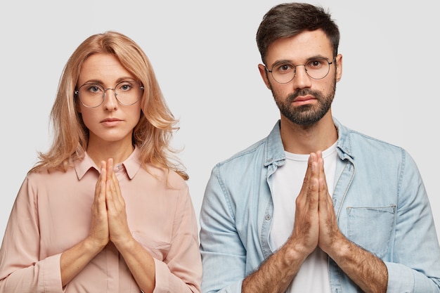 Free photo family couple stand in praying gesture, ask god to give them child. hopeful blonde female can`t get pregnant, believes in good fortune. attractive bearded young man wishes something desirable