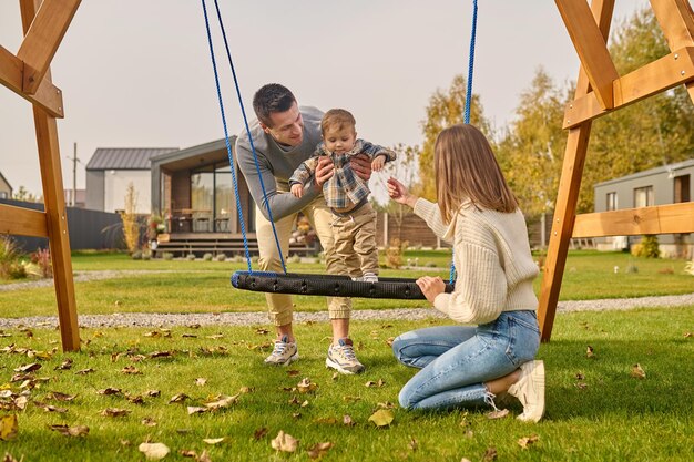 Family, leisure. Young adult smiling man supporting child on swing and woman crouching with outstretched hand spending leisure time together near country house on autumn day