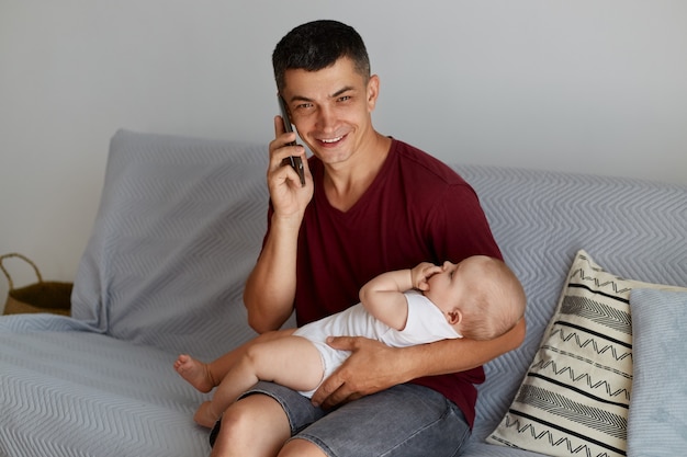 Family, parenthood and childhood. Portrait of happy smiling father with little baby boy or girl calling on smartphone at home while sitting on sofa in living room.