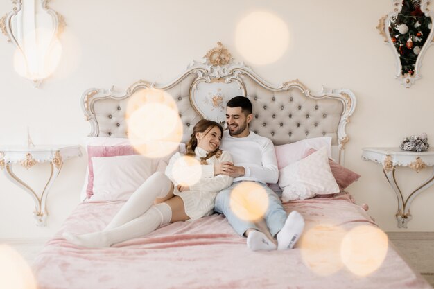 Family portrait. Man and woman relax on soft grey bad in a room with Christmas tree