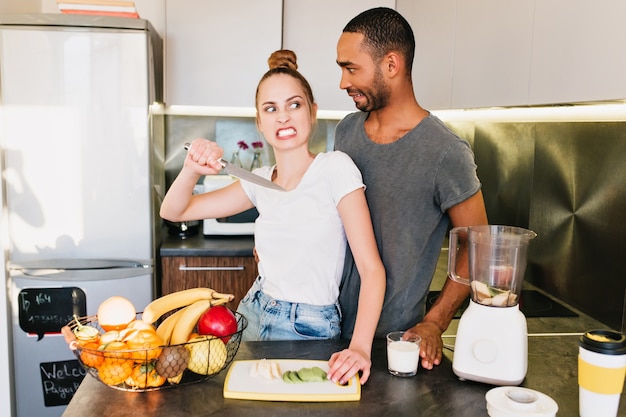 Family quarrel in the kitchen. Girl with angry face with a knife and a surprised guy. The husband and wife arguing, misunderstanding, broken relationships, irritated face, imitation quarrel.