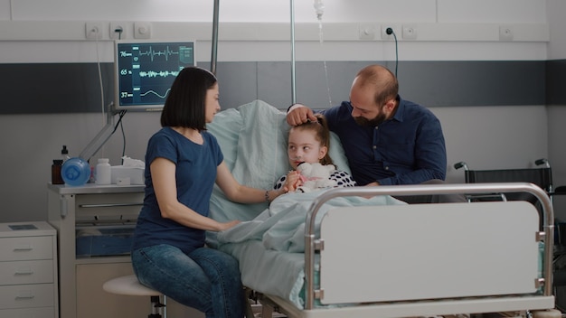 Free photo family sitting beside hospitalized sick daughter discussing medication recovery treatment during medicine examination in hospital ward. child resting in bed after suffering disease infection surgery