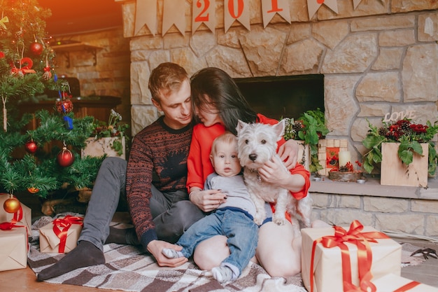 Free Photo family sitting in front of the fireplace with their baby and their dog and a christmas tree