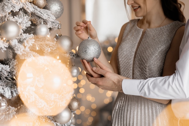 Free photo fancy dressed man and woman in silver gown hug each other tender standing before a christmas tree