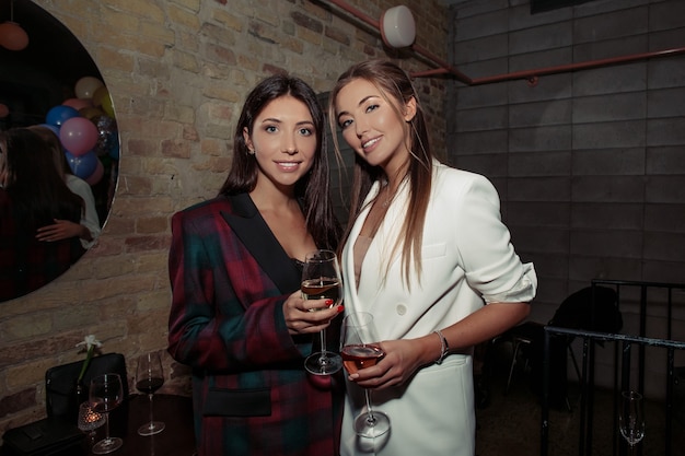 Fancy girls having fun on a party. Blonde wearing white jacket and accessories, natural makeup on the face and stylish hair. Brunette woman in a black fashionable dress.