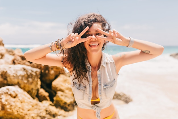 Fantastic joyful slim woman with wavy hair dressed in swimsuit and denim shirt on the beach