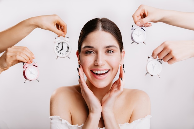 Fascinating girl without makeup, posing on isolated wall with alarm clocks.Brunette with green eyes smiles.