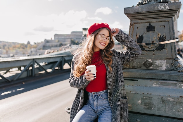 Free photo fascinating shy woman in glasses drinking coffee on bridge in windy day