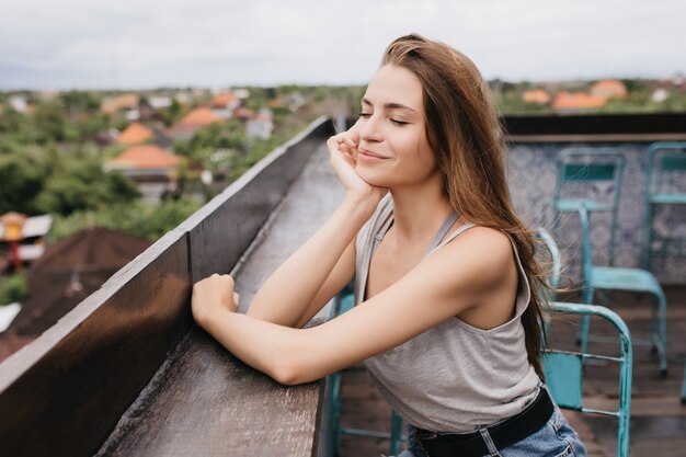 Fascinating young woman chilling at roof with eyes closed and romantic smile. Outdoor shot of girl in stylish clothes relaxing in spring day