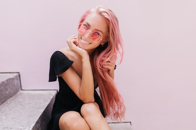 Free photo fascinating young woman in glamorous glasses smiling while sitting on steps.