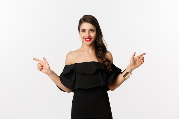 Fashion and beauty. Attractive woman in jewelry, makeup and black dress, laughing and pointing fingers sideways copy spaces offer, white background