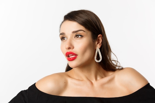 Free Photo fashion and beauty concept. close-up of luxury woman with red lips, earrings and black dress, looking away sensual, standing over white background