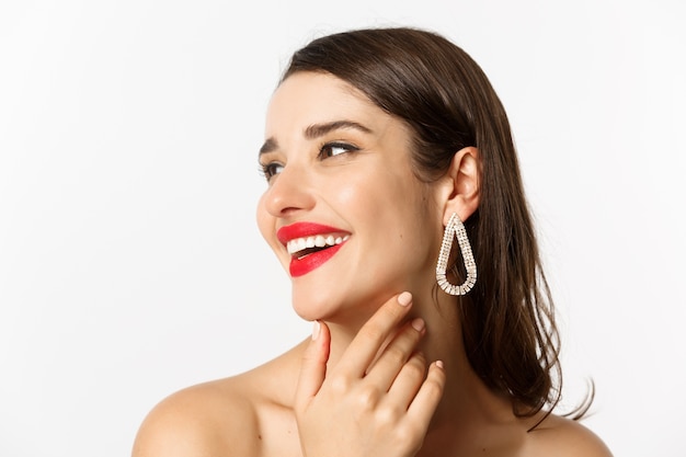 Fashion and beauty concept. Headshot of gorgeous brunette woman with red lipstick, earrings, laughing and looking left, standing over white background.