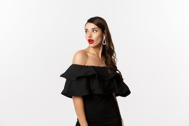 Fashion and beauty concept. Image of attractive young woman in black dress turn behind and looking at copy space, standing over white background