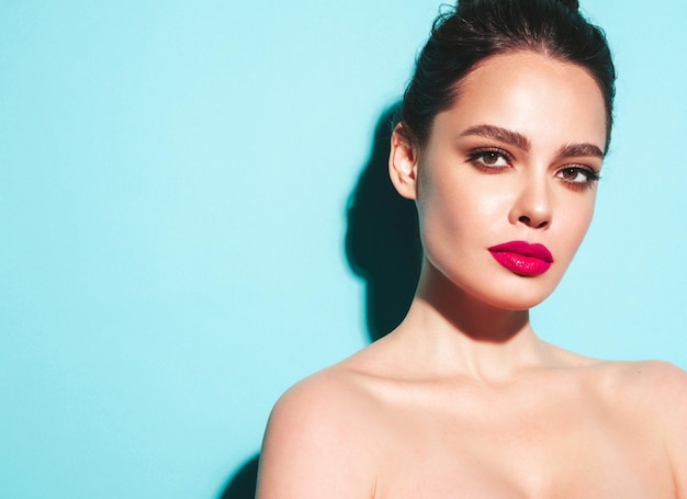 Fashion beauty portrait of young brunette woman with evening stylish makeup and perfect clean skin Sexy model with hair in a bun posing in studio near blue wall With pink bright natural lips