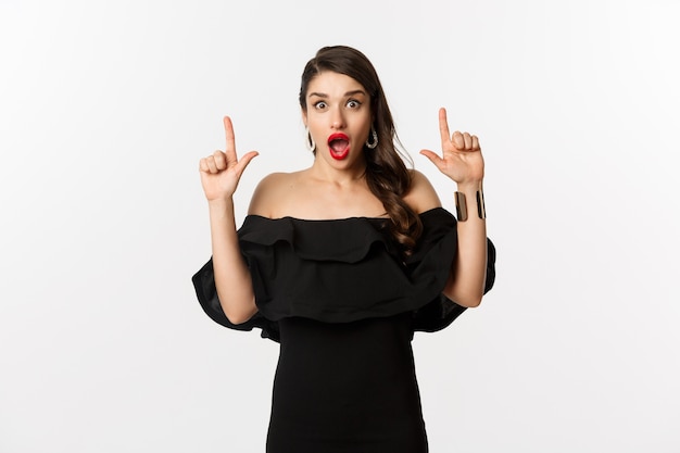 Fashion and beauty. Surprised woman in black dress pointing fingers up, showing banner, standing over white background.