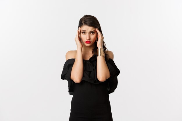 Fashion and beauty. Young modern woman in glamour dress, jewelry and makeup, touching head and looking exhausted, feeling dizzy, standing over white background.