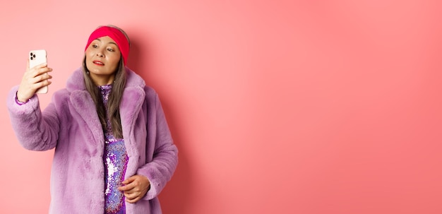 Free photo fashion concept stylish asian senior female taking selfie on smartphone posing in purple faux fur co