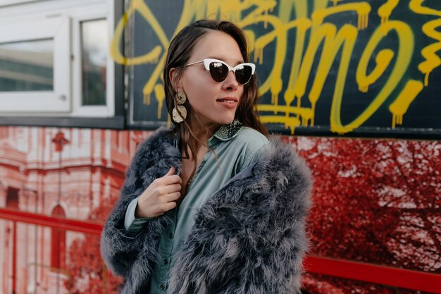 Fashion glamour woman in trendy fur coat and glasses with gold jewelry posing on city