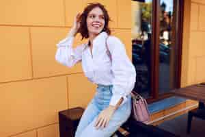 Free photo fashion image of pretty brunette girl with short hairstyle in stylish casual white blouse and jeans. black leather boots on heels . girl sitting near modern cafe with yellow walls.