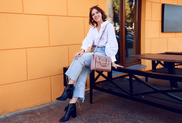 Free Photo fashion image of pretty brunette girl with short hairstyle in stylish casual white blouse and jeans. black leather boots on heels . girl sitting near modern cafe with yellow walls.