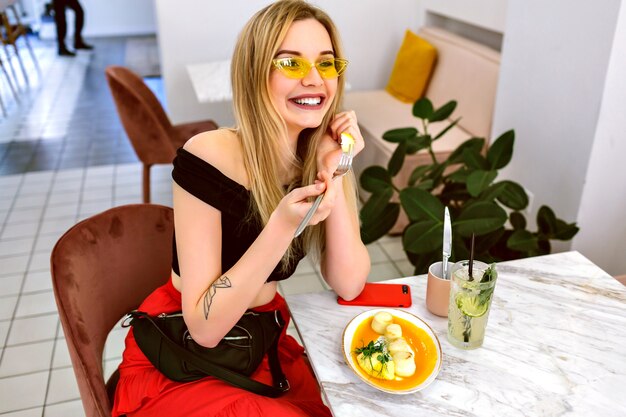 Fashion indoor portrait of young trendy hipster blonde woman posing at modern hipster cafe, morning breakfast time.