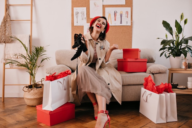 Free photo fashion lady in red beret and beige coat laughing and holding black sandals. joyful young girl with dark hair enjoys shopping.