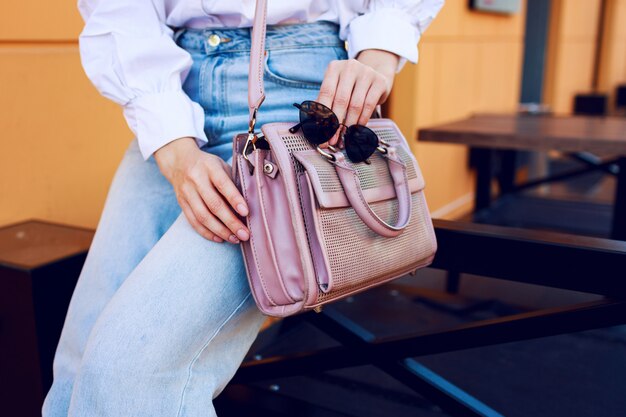 Fashion objects. WomanÃÂ¢ÃÂÃÂs hands with bag and sunglasses. Fashionable girl sitting outdoor . Stylish jeans.