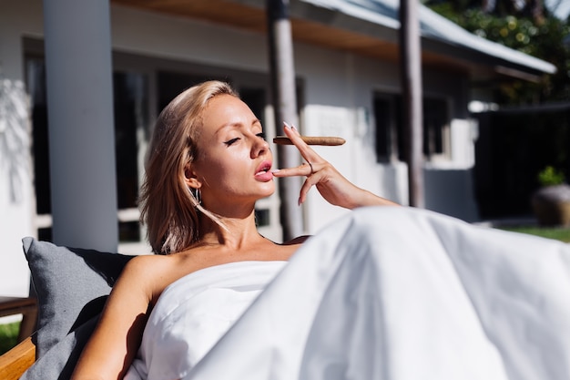 Free photo fashion outdoor portrait of naked woman sits on sofa covering herself with blanket holding cigar,