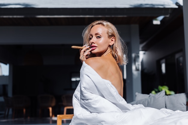 Fashion outdoor portrait of naked woman sits on sofa covering herself with blanket holding cigar