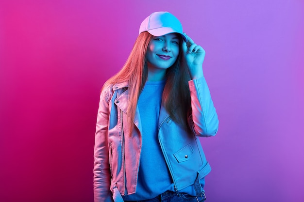 Free photo fashion portrait of confident woman standing against pink neon wall and touching visor of her cap, wearing leather jacket, looking at camera